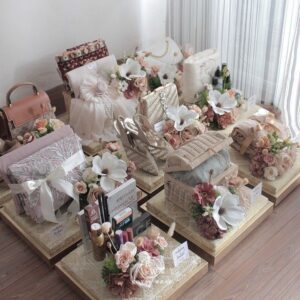 Elegant wedding gift hampers arranged on decorative trays, adorned with pastel flowers, luxury accessories, makeup, and clothing, placed near a window with sheer curtains.