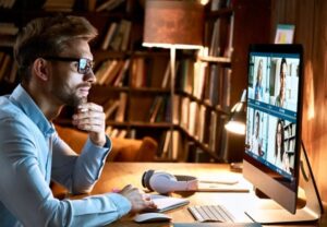 Man wearing glasses participating in a virtual meeting on a desktop computer in a cozy, book-filled home office.